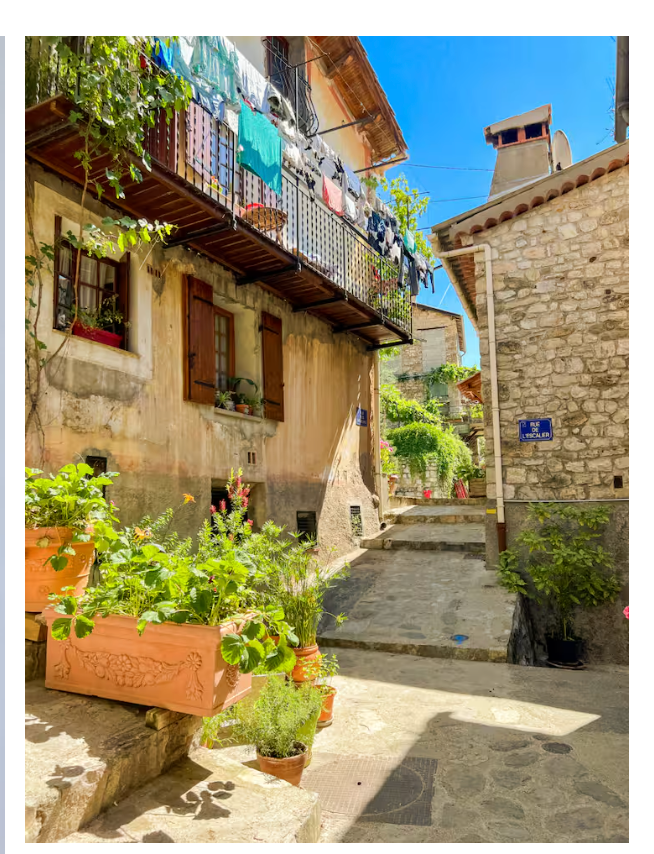 Rue paisible du village de Touët-sur-Var, maisons en pierre, plantes et balcons fleuris sous le soleil.